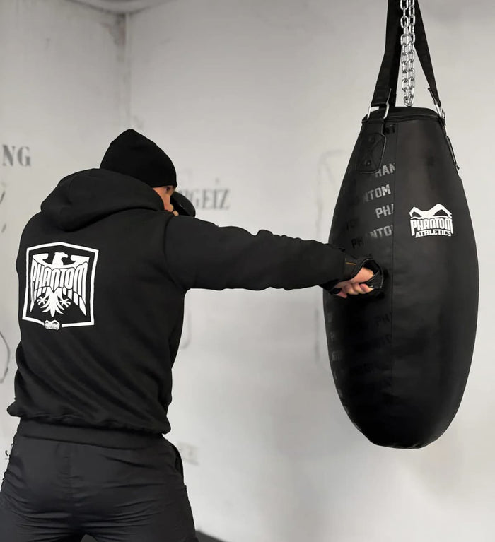 Kampfsport-Athlet mit Phantom Athletics Hoodie Germany, MMA-Handschuhen, Trainingshose und Mütze beim Boxsacktraining am Boxsack Bodyshot