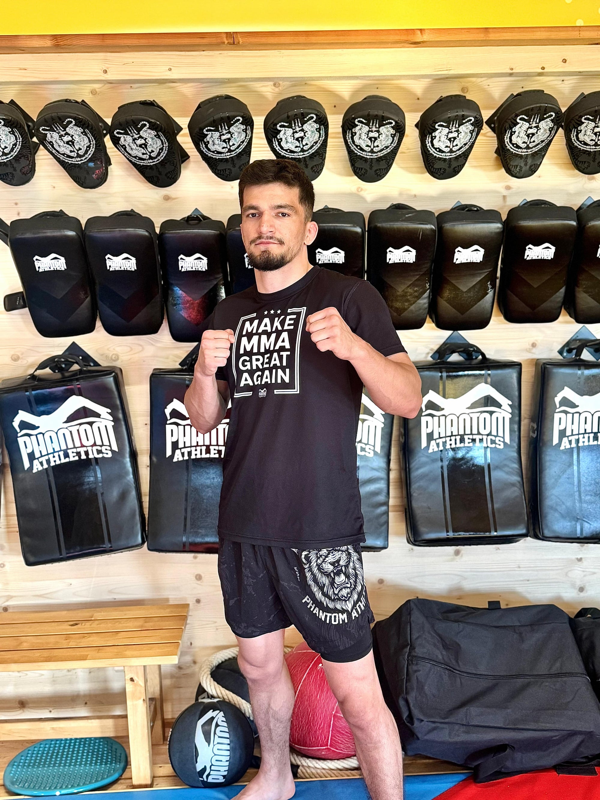 MMA-Fighter Rafiew "RAFAJUNIOR" Komron mit Phantom Athletics T-Shirt, Trainingshorts und Pratzen im Hintergrund