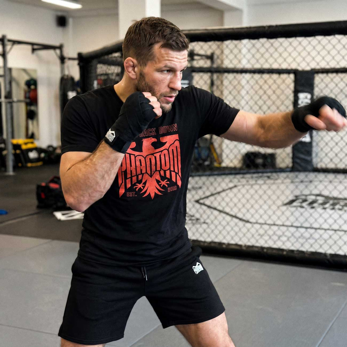 Das schwarze Phantom Athletics GERMANY NEVER BACK DOWN T-Shirt wurde für Kampfsportler entwickelt, die einen markanten Fighter-Look mit hohem Tragekomfort verbinden möchten. Der rote Eagle-Print macht das Shirt zum Blickfang beim Training, im Gym und im Alltag.