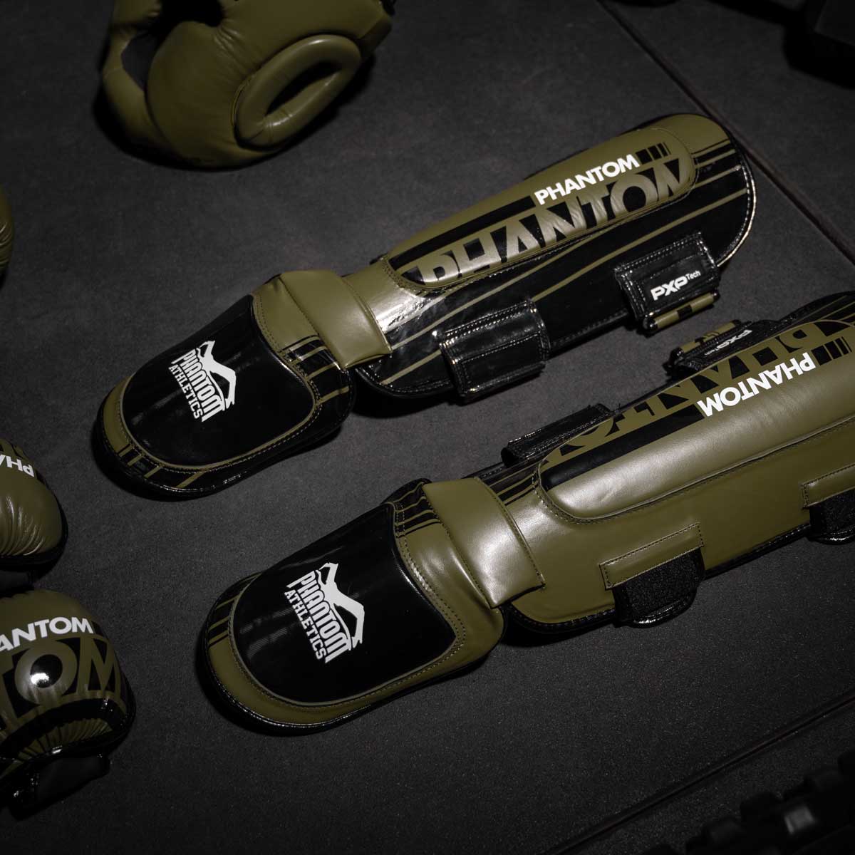 Die army grünen Phantom Apex Hybrid Schienbeinschoner am Gymboden neben dem Kopfschutz und MMA Handschuhen.