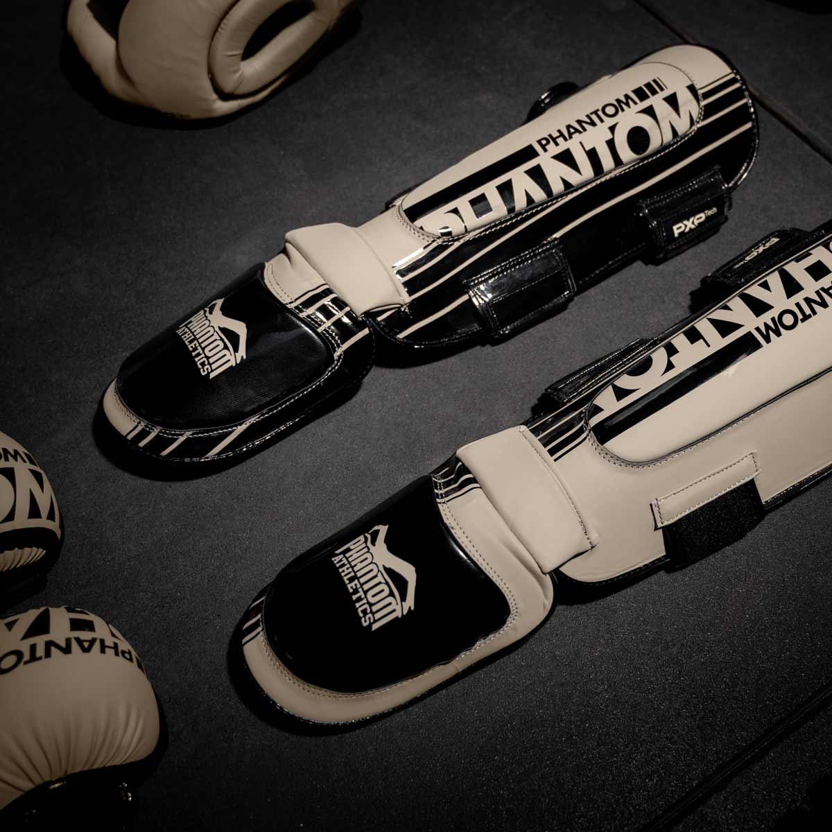 Die sandfarbenen Phantom Apex Hybrid Schienbeinschoner am Gymboden neben dem Kopfschutz und MMA Handschuhen.
