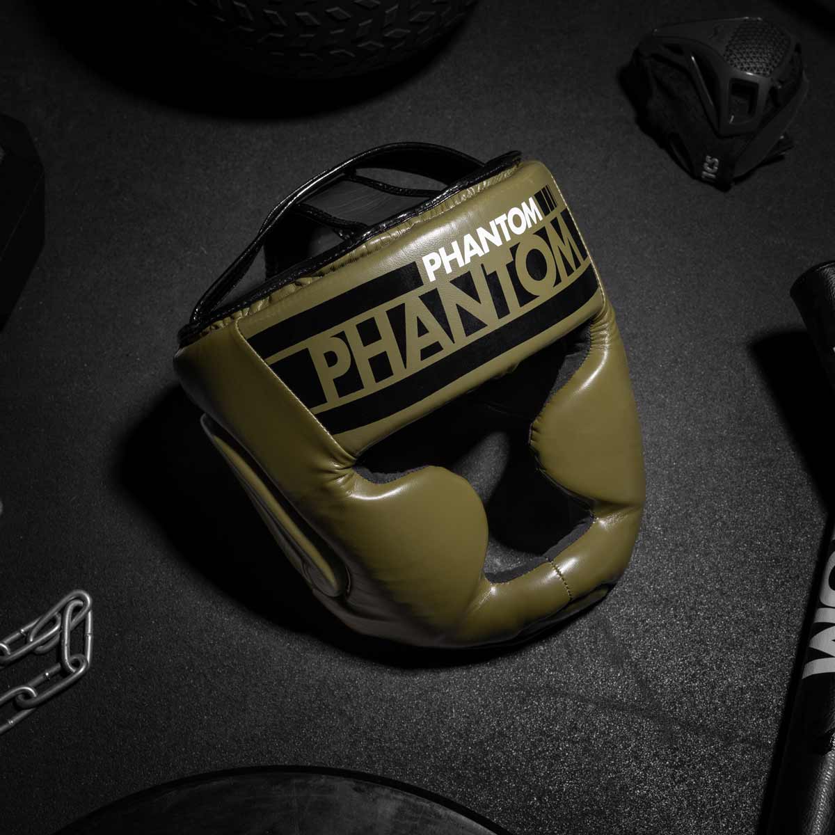 Der Phantom APEX Full Face Kopfschutz in Army Grün am Gymboden neben der Phantom Trainingsmaske
