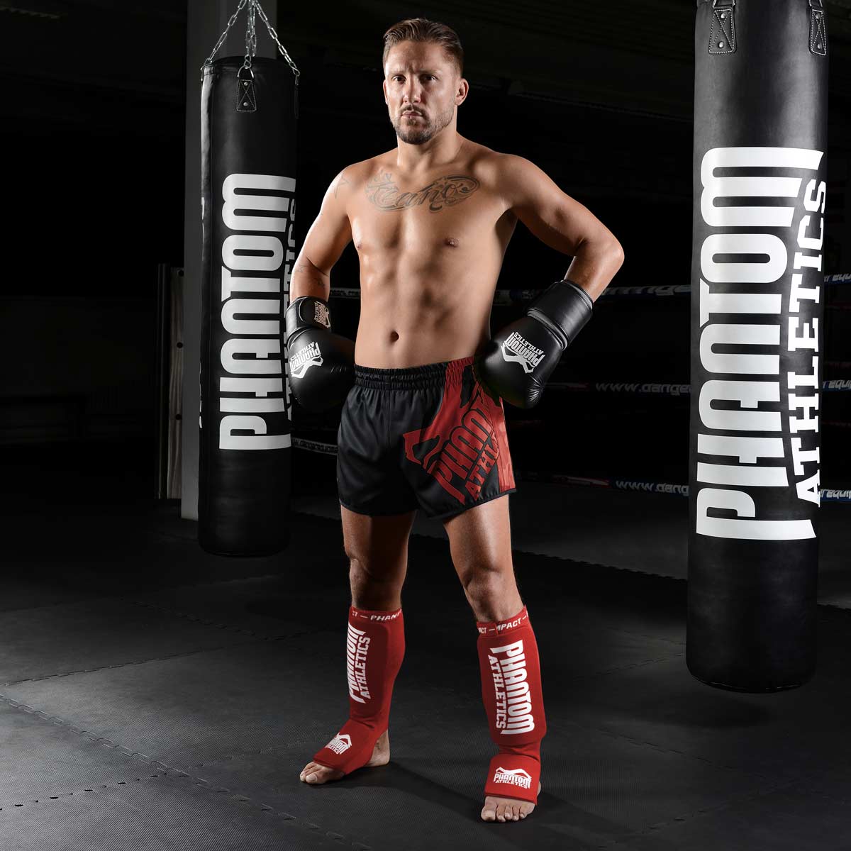 Kickboxer mit den Phantom Impact Kampfsport Schienbeinschonern in Rot im Gym vor den Boxsäcken.